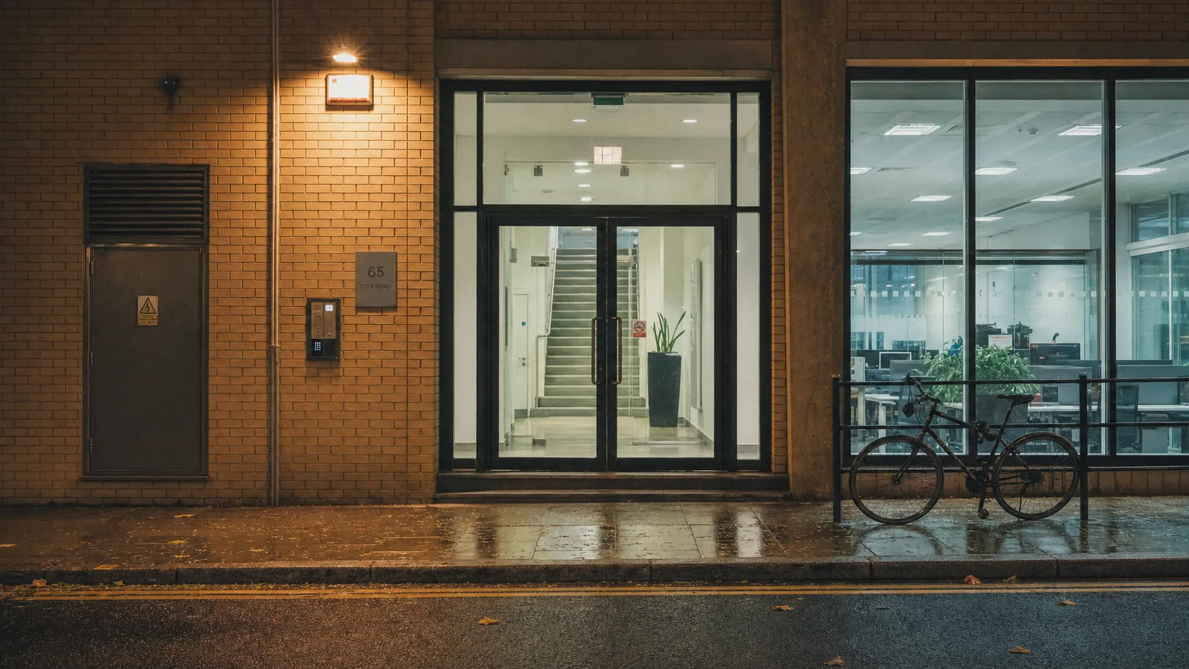 A plain London office doorway on City Road at night, entryphone panel beside the glass, a lit stairwell visible inside and a bicycle locked at the kerb.