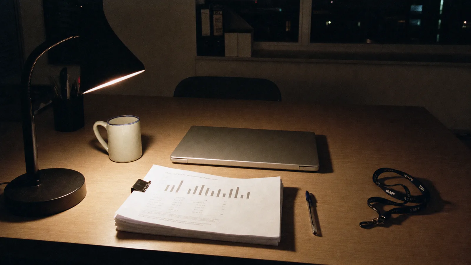 A plain office desk at night lit by a single lamp: a closed laptop, a clipped stack of printed documents, a pen and a staff lanyard.