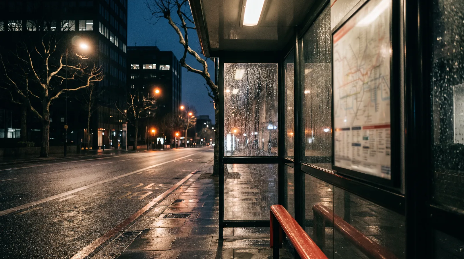 An empty London bus shelter at night after rain, the timetable panel lit inside and the wet road reflecting street light.