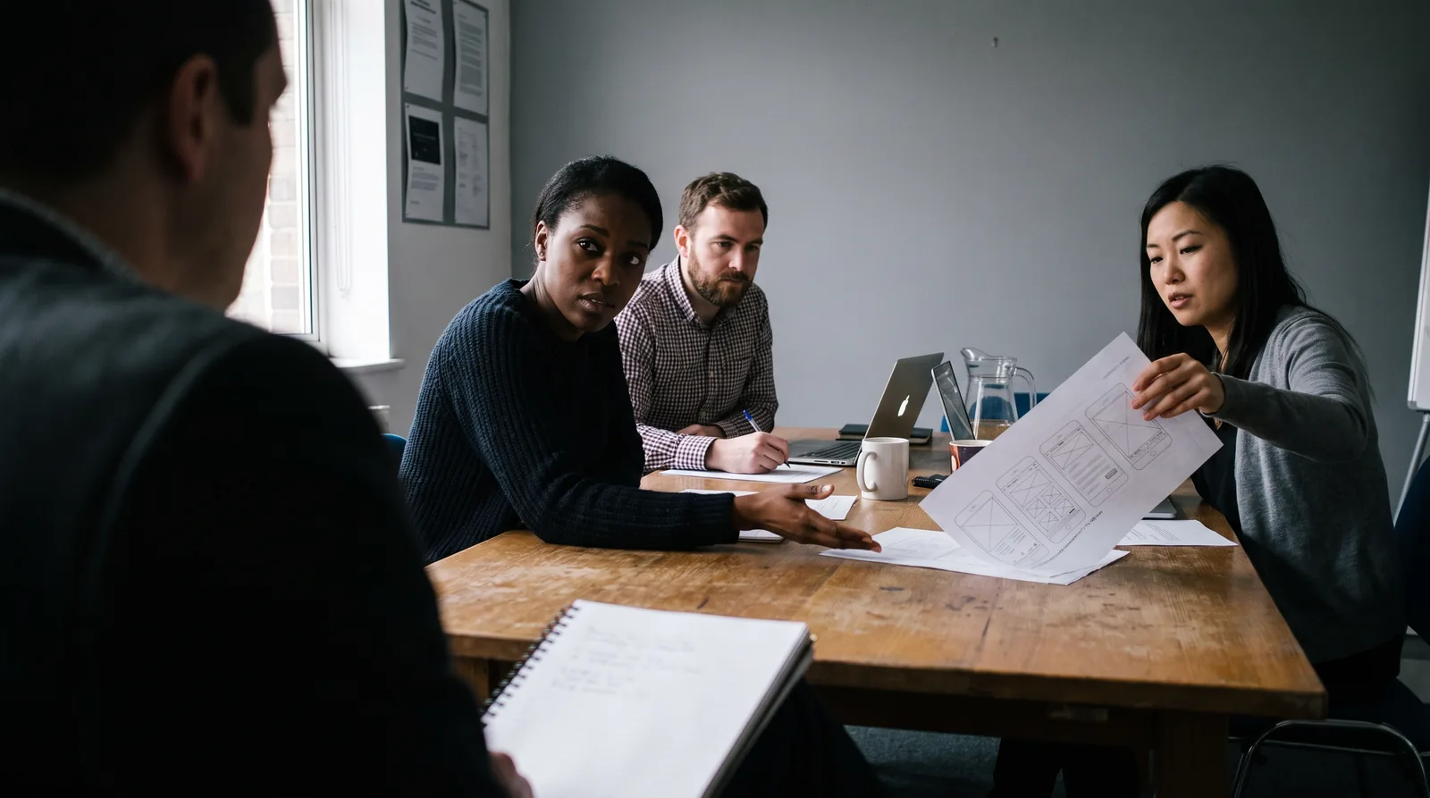 Three of the team across a meeting table from the viewer, one explaining with an open hand, one taking notes, one turning a printed layout round to face the client.